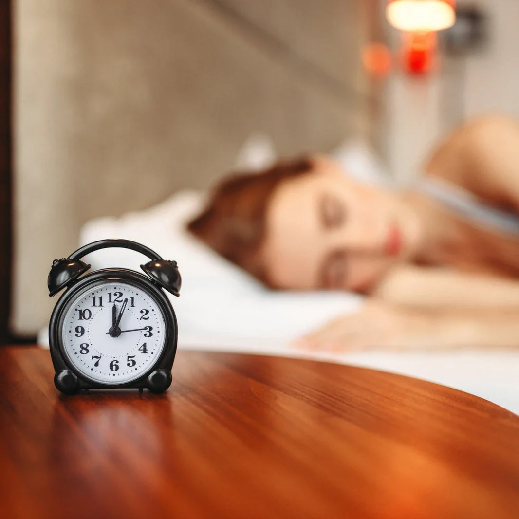 Analog black alarm clock at 3 AM on bedside table, foreground focus, with a woman peacefully sleeping in bed in soft blur behind, symbolizing good sleep.