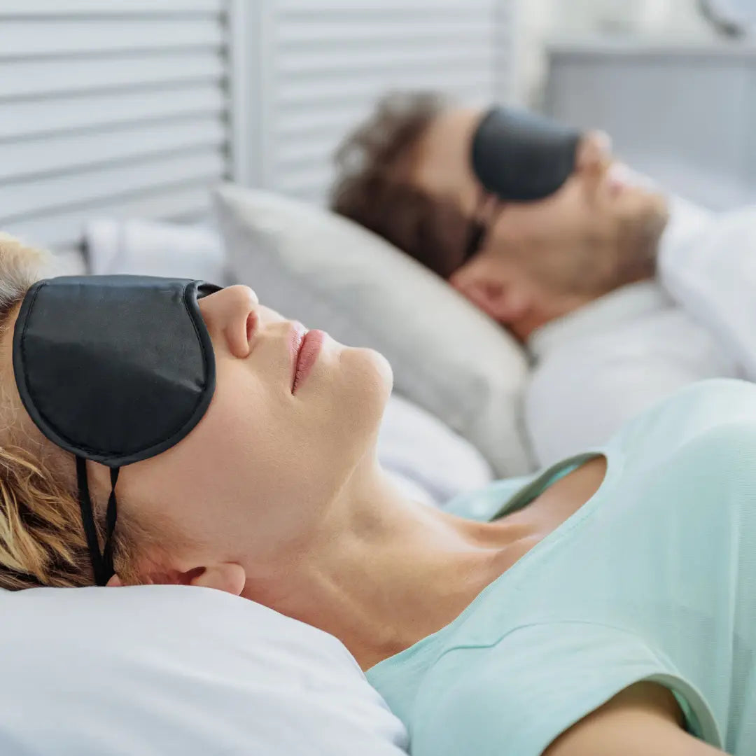 Couple sleeping soundly in bed wearing blackout eye masks, eyes closed in relaxation on pillows in cozy bedroom setting.