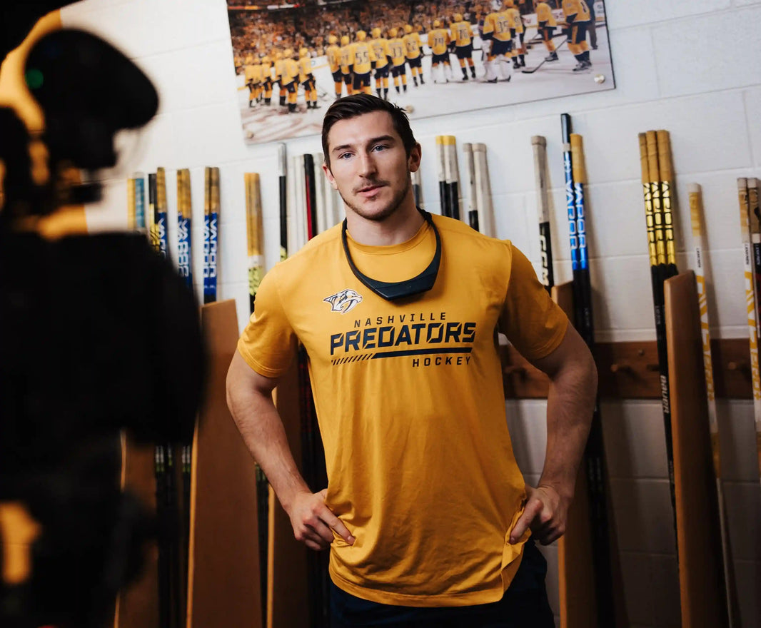 Nashville Predators player in yellow team shirt and Hapbee neckband in locker room with hockey sticks and team poster, promoting wellness and recovery tech.