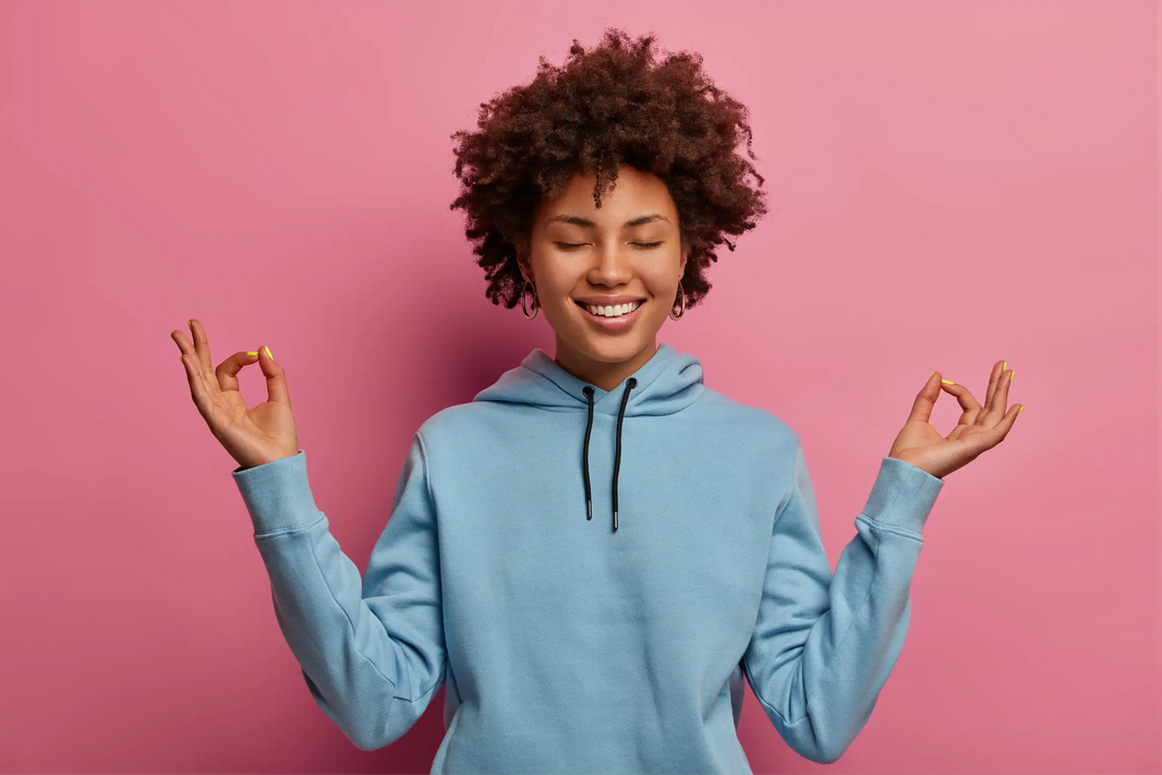 Smiling Black woman with afro in blue hoodie meditating with eyes closed and zen hand gesture against pink background.