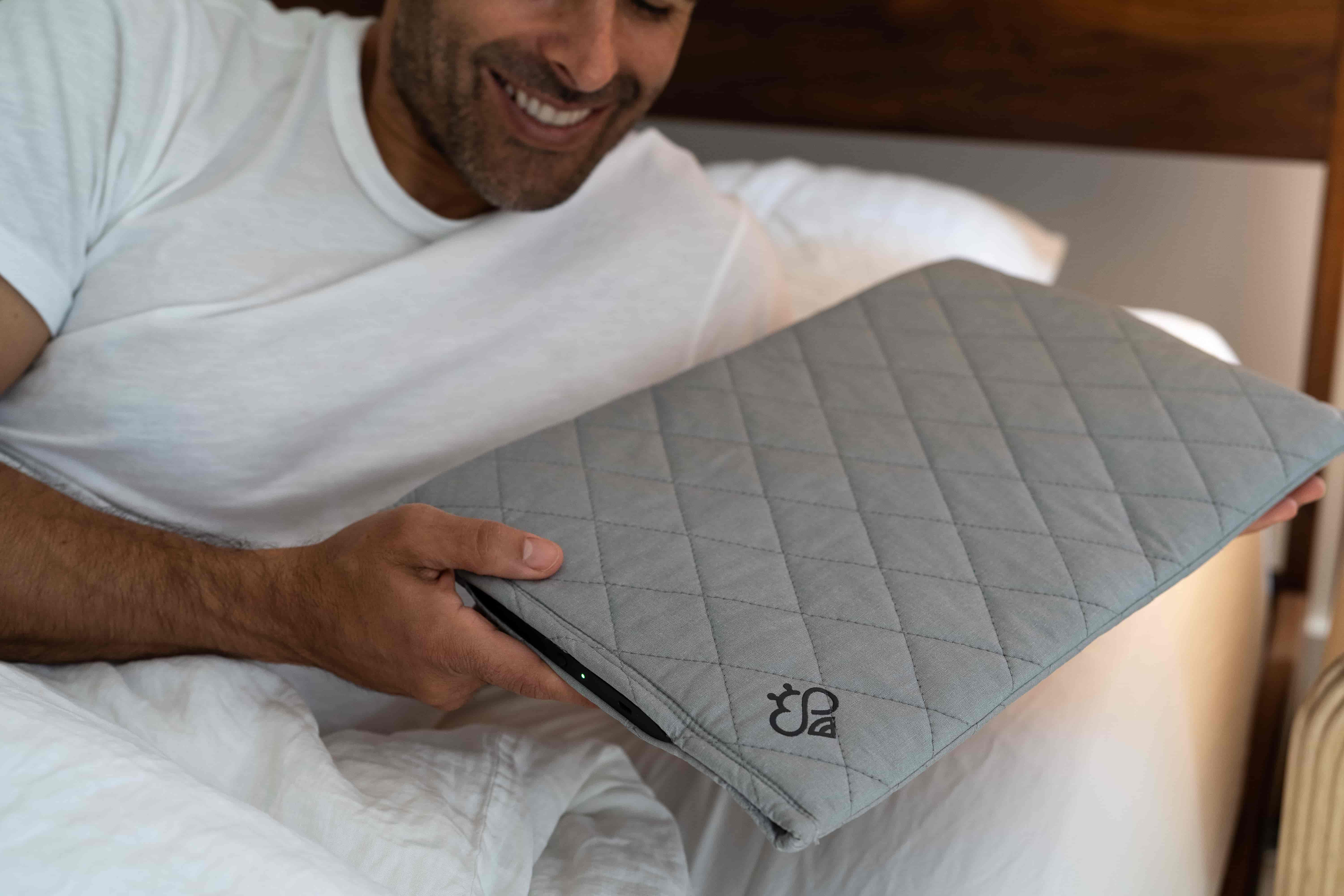 Man holding a gray quilted Hapbee Sleep-Pad with a brand logo on a bed.