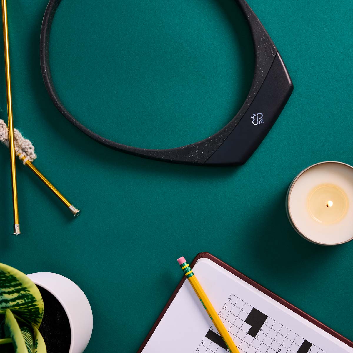 Hapbee black neckband on teal backdrop styled with crossword puzzle, pencil, lit candle, gold knitting needles, and succulent plant for relaxation theme.