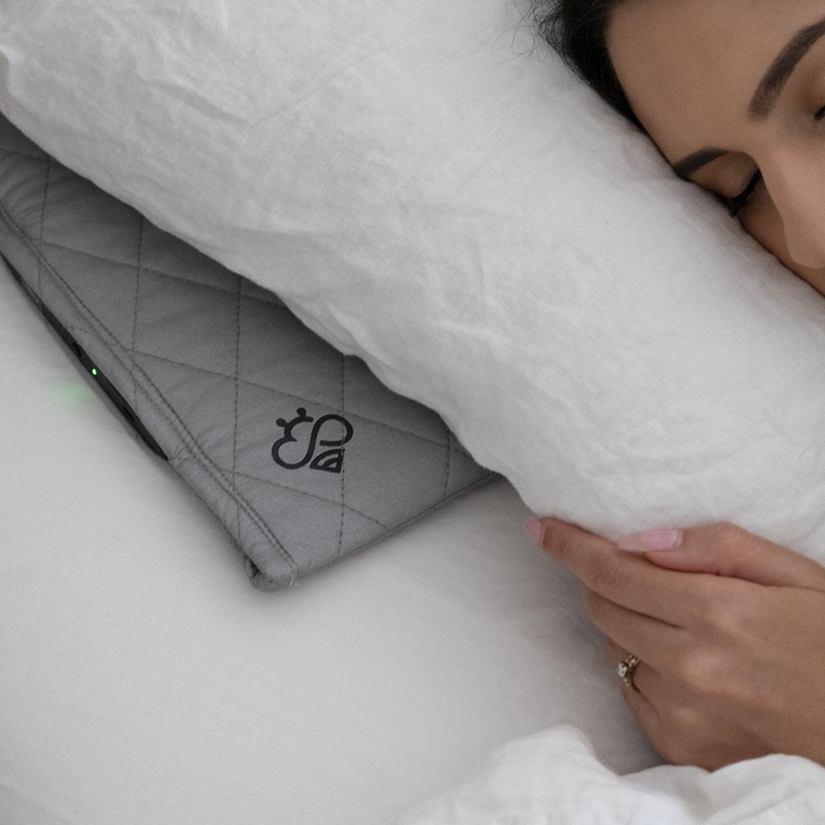 Peaceful woman sleeping with Hapbee Sleep Pad under white pillow, gray quilted pad showing logo and green light, cozy rest scene.
